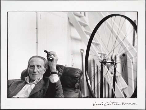 The artist sits in a leather chair wearing a suit jacket and holding a cigar. Next to him is a large bicycle wheel attached to a stool.