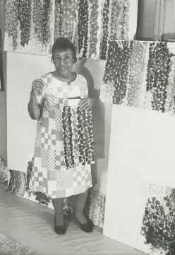 A black and white photo of the artist in her studio. She wears a patchwork style dress and stands holding one of her paintings.