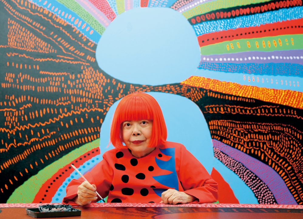 Portrait of Yayoi Kusama infront of a colorful painting.