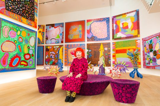 The artist wears a pink and black polka dotted dress. She is sitting on a bench that matches the pattern of her dress. There are large square paintings on the walls around her. The paintings are full of brightly colored shapes.