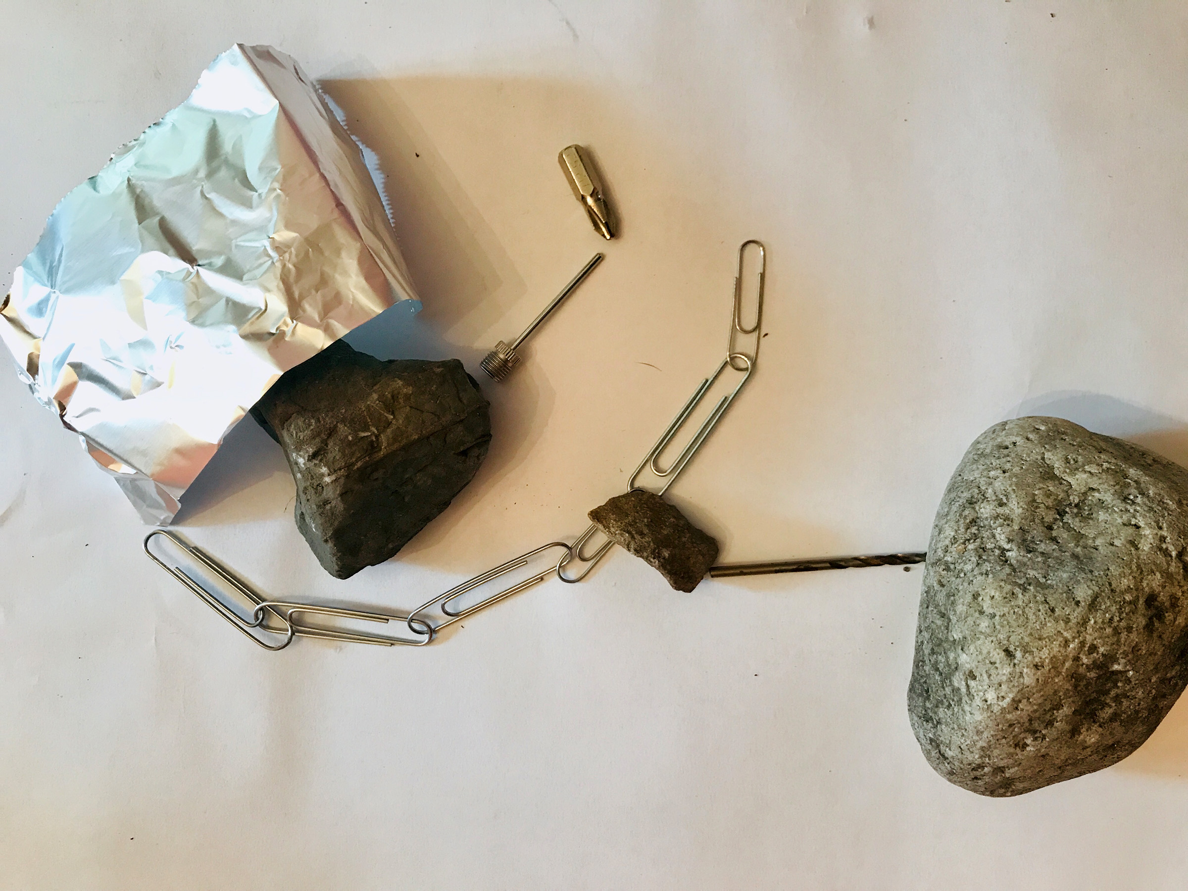 Next to a drill bit and inflation needle, a rock comes out of a small foil tunnel. There is a chain of paperclips in a half circle by the foil tunnel. A larger grey and tan rock is near the edge of the image.