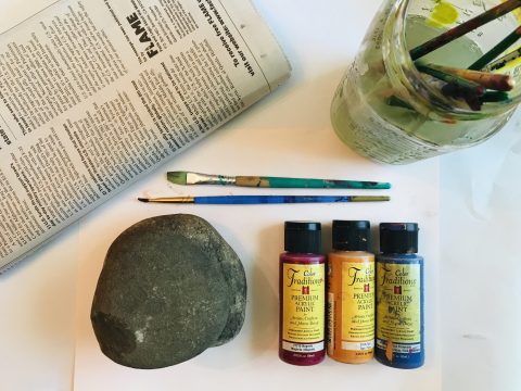 Folded newspaper, two paint brushes, a clean rock, three small bottles of paint, and a glass jar with paint brushes in dirty water