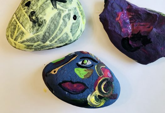 Three rocks, one painted yellow with black lines, one purple with pink lines, one blue with multicolored, misaligned facial features