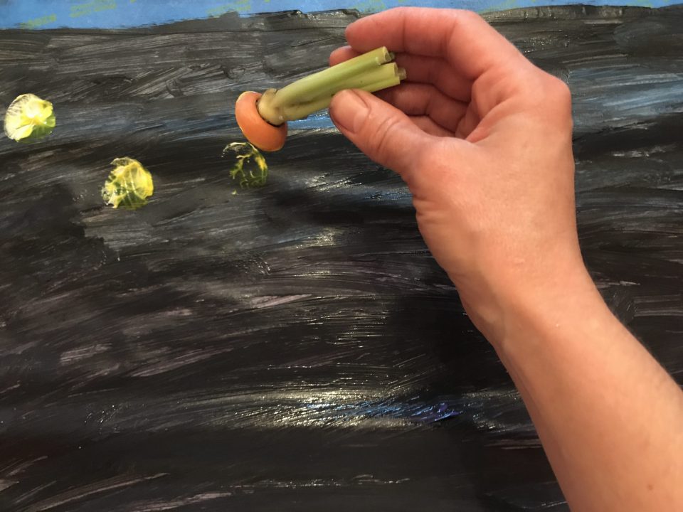 Hand using a sliced carrot to paint yellow dots on a black background