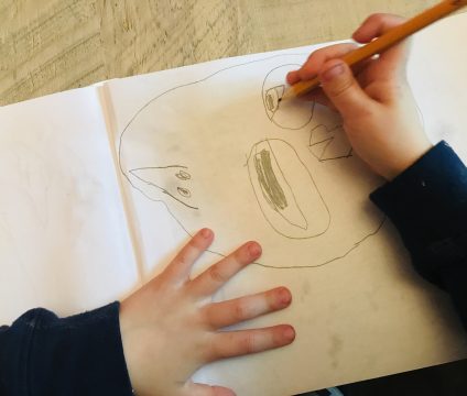 Pencil sketch of a face with two different sized eyes aligned vertically in the center, pointed nose on the left, mouth on the right. A child’s hand is drawing the eye to the far right.