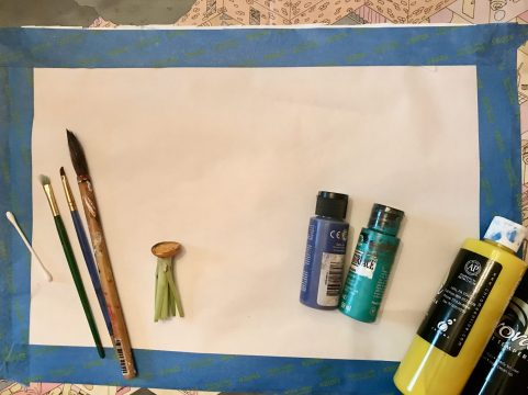 Paintbrushes, a cotton swab, a sliced carrot, and bottles of paint lie on a large piece of paper outlined with painter’s tape