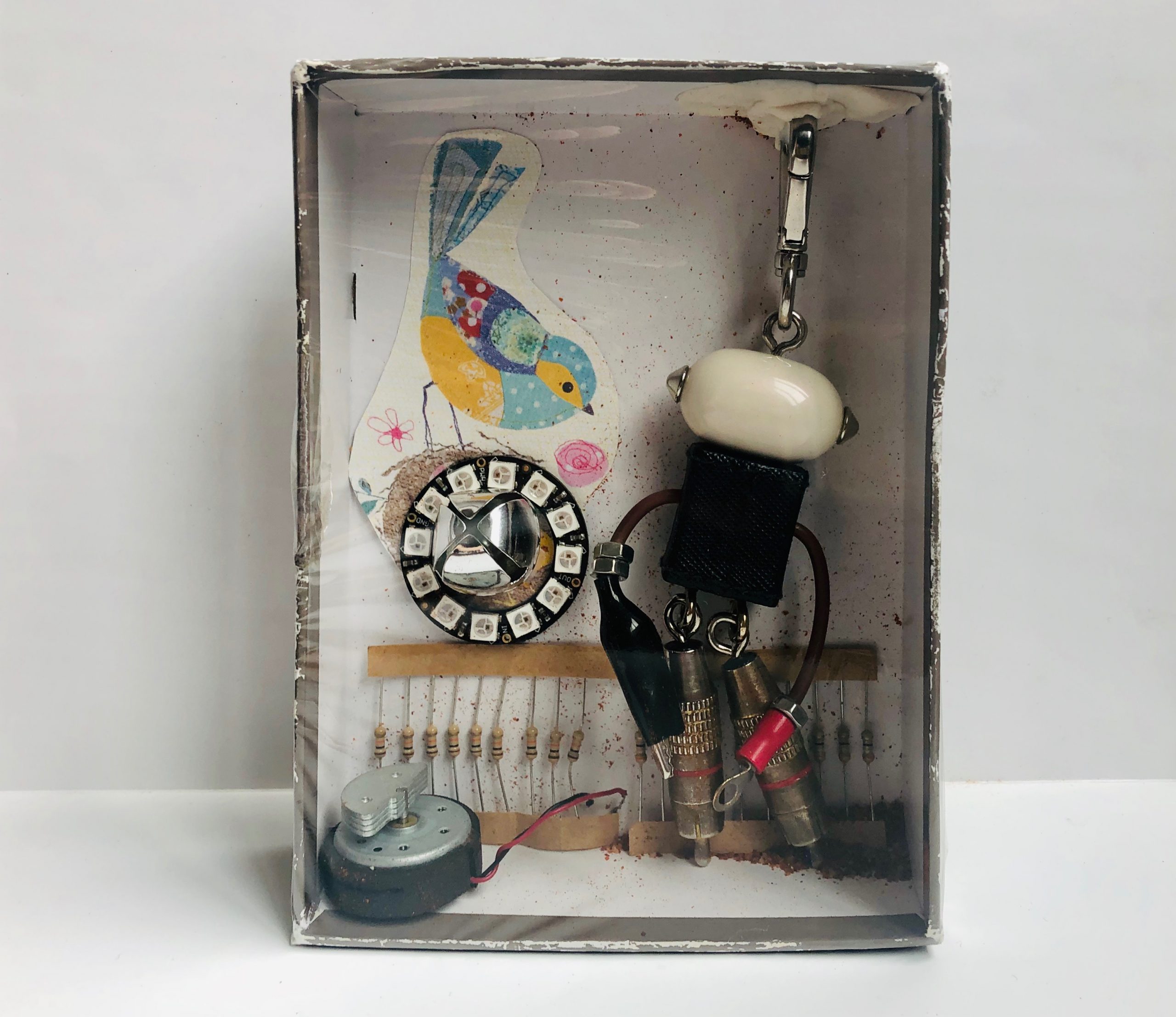 Final product of a small cardboard box covered with plastic wrap containing keychain, paper cutout of bird, jingle bell, resistors, and other found objects