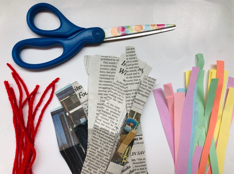 Pair of scissors lying above red yarn, newspaper, and multicolored paper all cut into strips