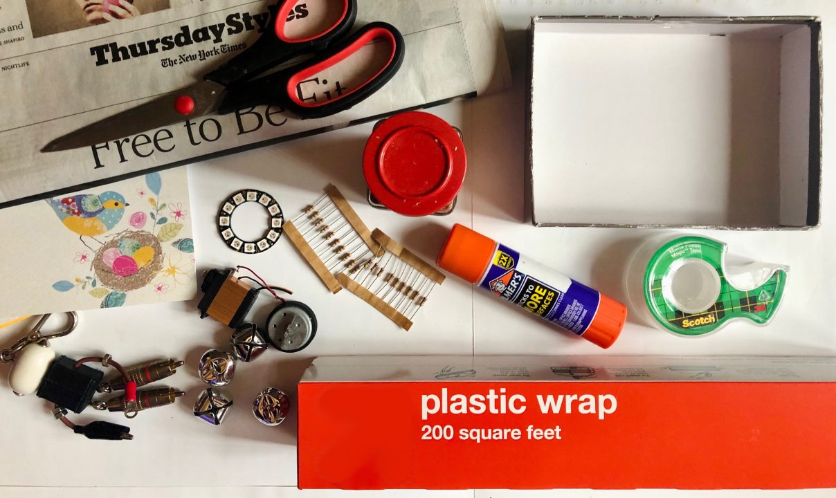 Pair of scissors on a newspaper, jar lid, glue stick, roll of tape, plastic wrap, four jingle bells, keychain, resistors, and more found objects