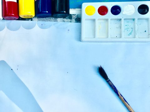 Overhead view of blank paper with paintbrush lying on it. Bottles of paint and a palette are at the top of the frame.