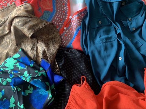 Close-up of a pile of clothing including a red patterned blouse, tan scarf, black scarf with blue and green flowers, and plain red blouse.