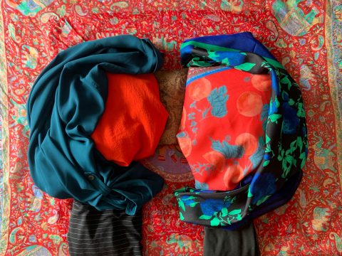 Blue shirt and black scarf are added to create the shape of two figures in profile facing each other, a hand reaches to pinch the fabric of one figure into a nose.
