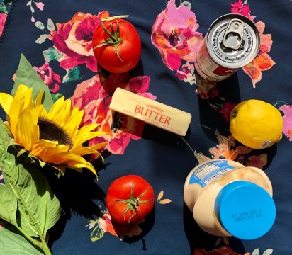 An overhead view of two tomatoes, a stick of butter, can of soup, lemon, bottle of mayo, and sunflower arranged on a piece of floral fabric.