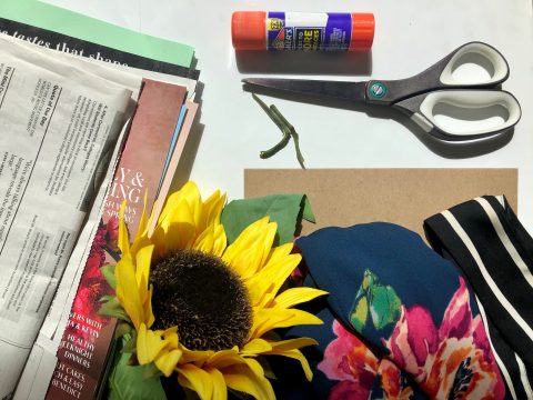 An arrangement of newspapers, multicolored paper, glue stick, pair of scissors, sunflower, floral fabric, and black fabric with white stripes.