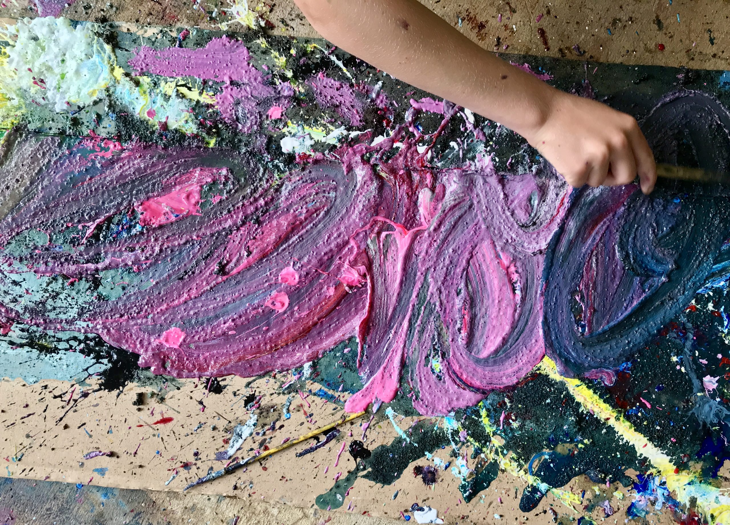 Cardboard surface covered with multicolored paint, a hand reaches to smear the paint with a brush.