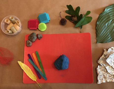 Rocks, popsicle sticks, a feather, and playdough lie on a red mat. Leaves, netting, tin foil, and dry pasta surround the mat.