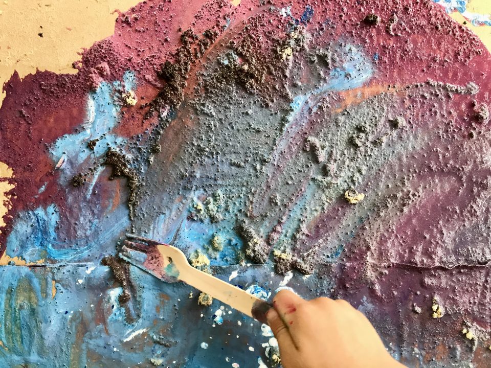 Close-up of a surface covered with a mixture of paint and a gritty material, with a hand using a fork to spread the paint.