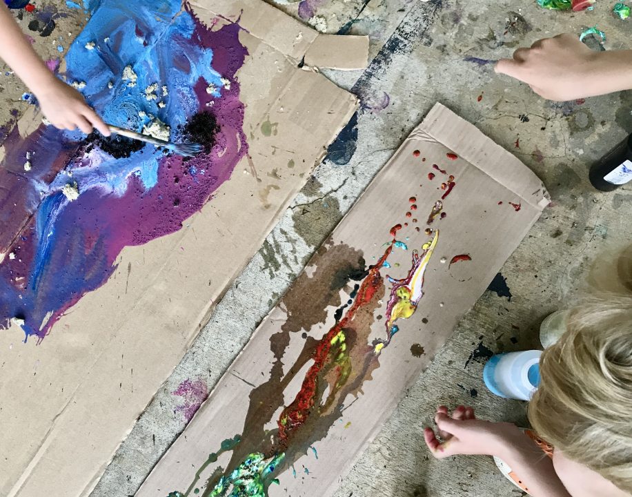 People leaning over two large pieces of cardboard, dripping and splashing paint onto the surface