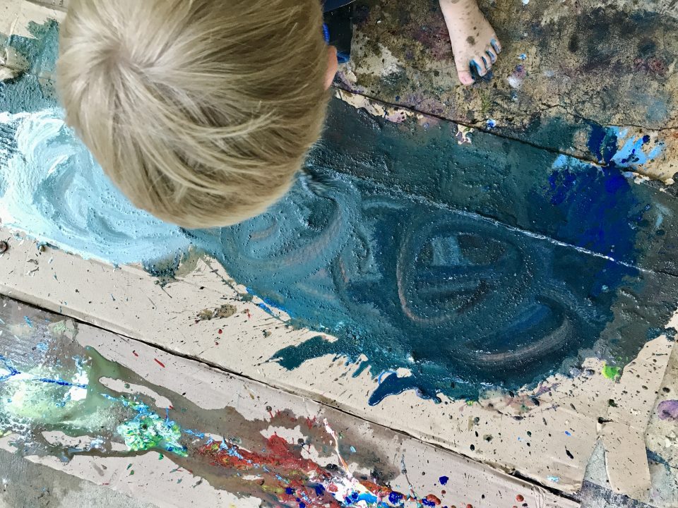 Overhead view of a child walking on a large piece of cardboard that is covered with splatters and smears of paint.