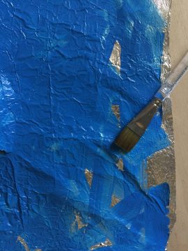 A paint brush rests on a rectangular piece of aluminum foil. The foil is mostly painted bright blue; a few sections of bare foil are exposed.