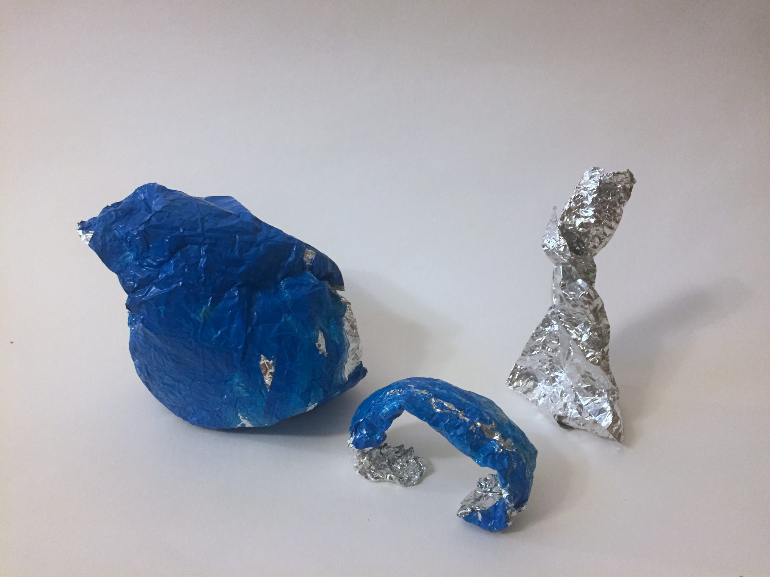 Three aluminum foil sculptures in a row. From left to right: a blue bird-like shape, a blue arch, and an unpainted triangle with a twisted top.