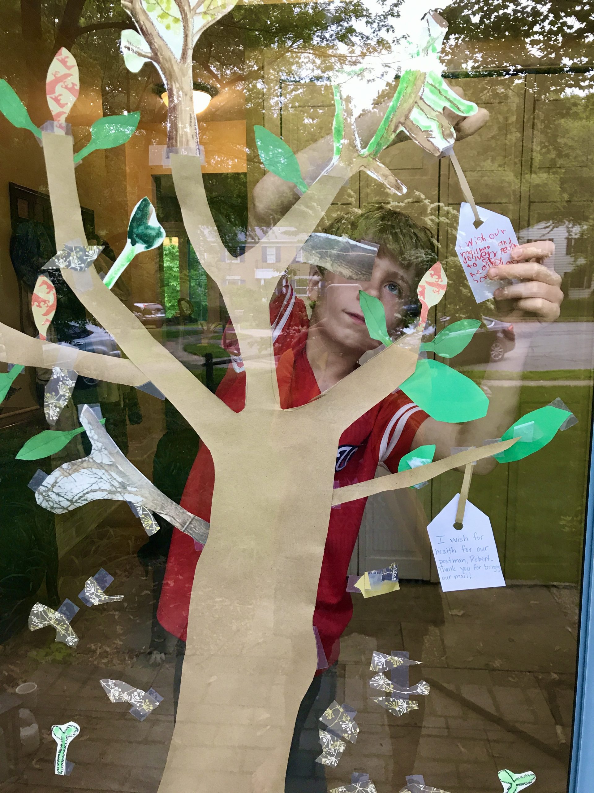 Exterior view of a paper tree with green paper leaves taped to a window, a boy reaches to hang a white tag on one branch