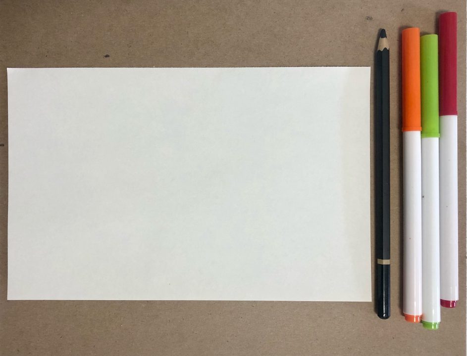 A piece of paper rests on a wood table. A pencil and three markers sit beside the paper.