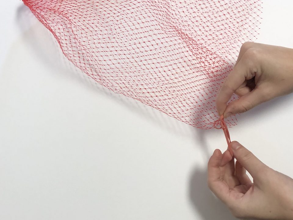 A hand ties a rubber band between the webbing of a red produce net.