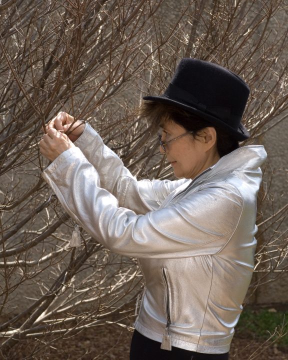 The artist in profile wearing a black hat and silver jacket hangs a tag on a bare tree