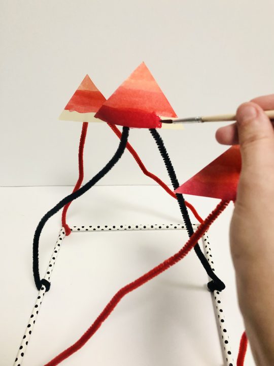 A hand holding a paint brush finishes a line of dark red across the bottom of a triangle. The triangle is suspended in the air by pipe cleaners.