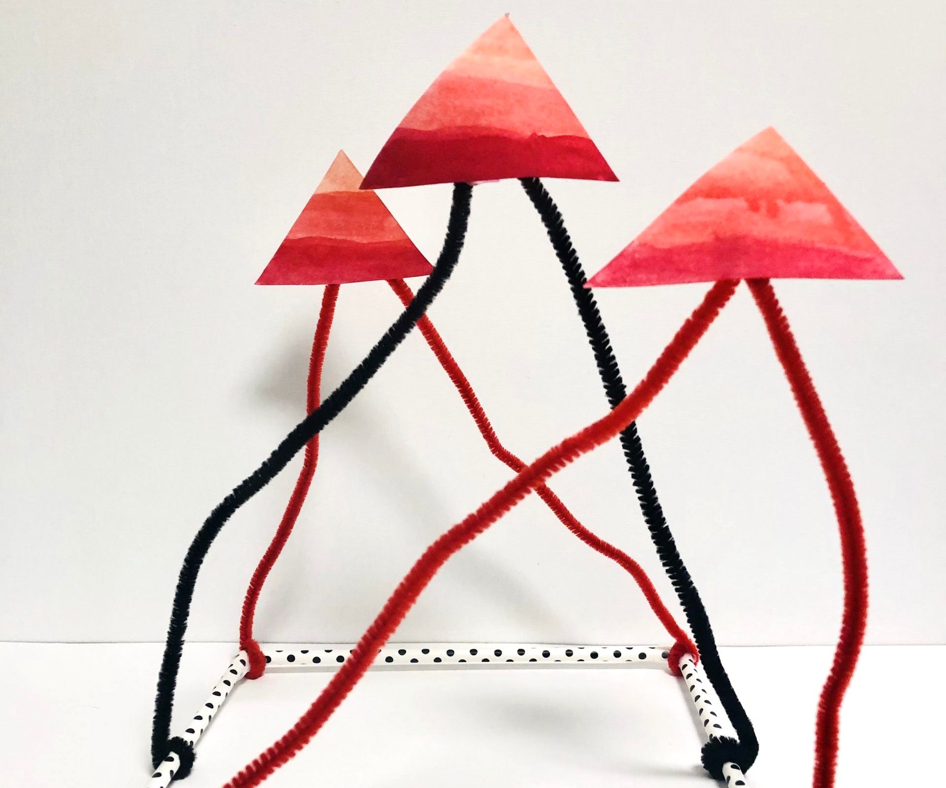 Red, black, and white structure. The square base is made from white polka-dot tubes. Pipe cleaners extend upward displaying three red triangles.