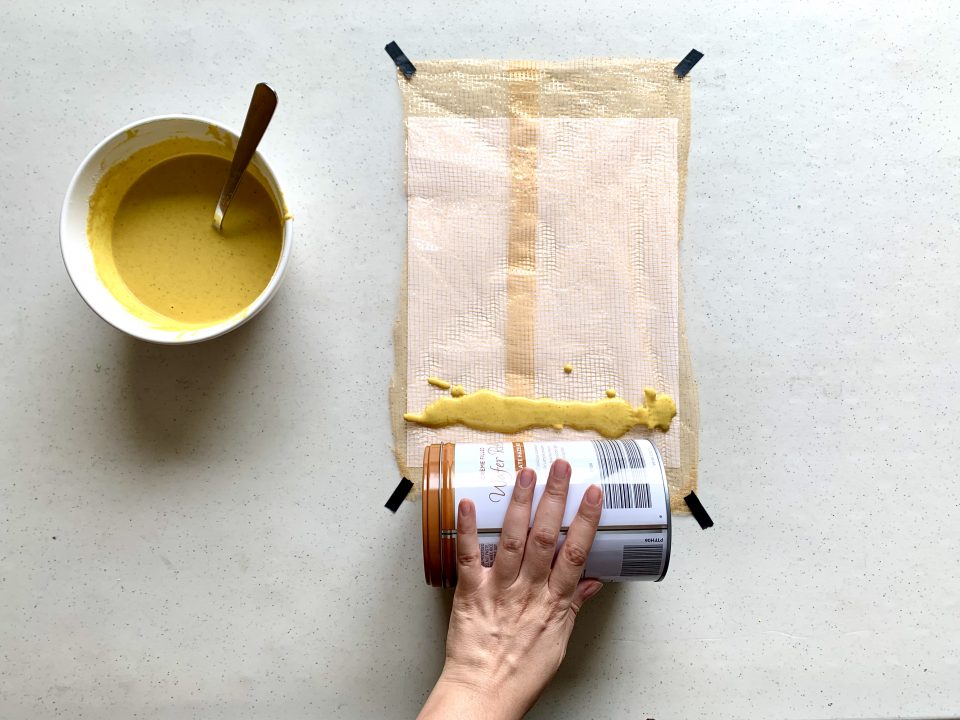 A paper covered with a net is taped to a table. A hand holds a tin can and prepares to roll the can over a line of yellow paint that’s drizzled atop the net.