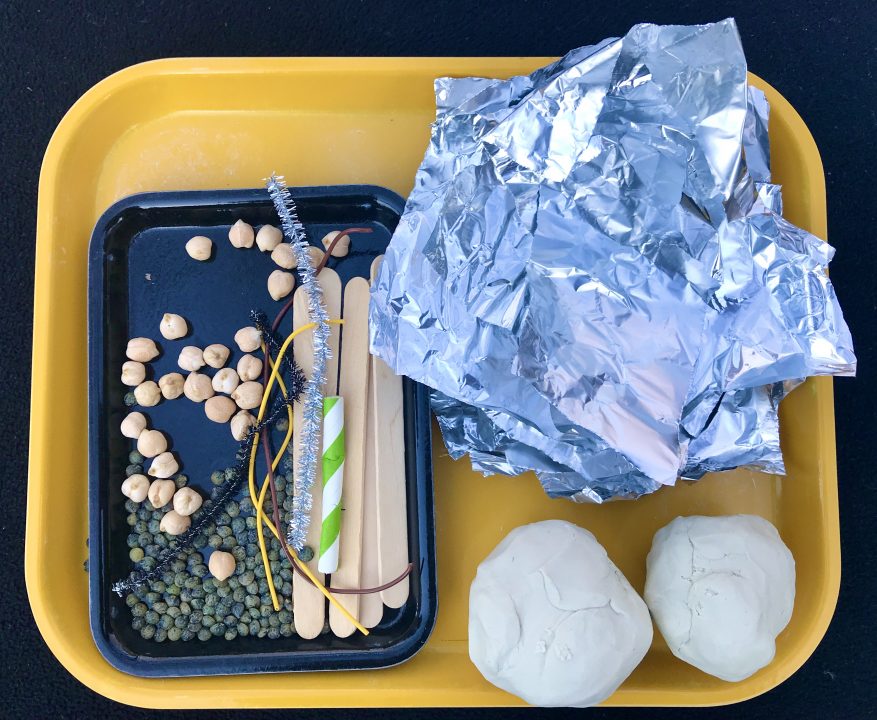 A yellow tray holds a stack of aluminum foil, two balls of white clay and a small black tray with dry beans and popsicle sticks.