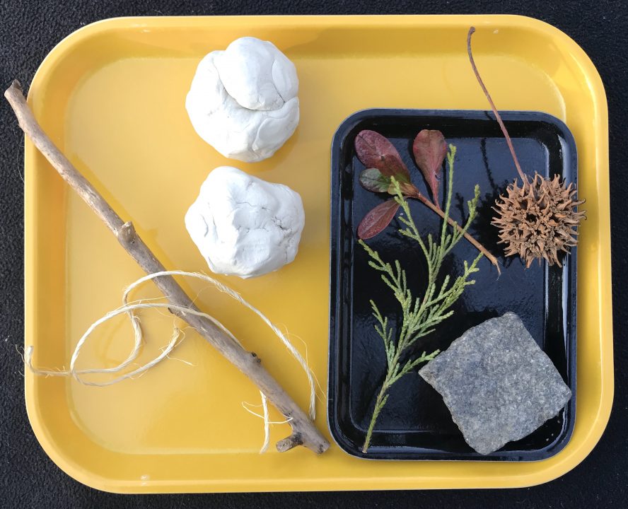 A yellow tray holds two balls of clay, a straight stick, a strand of twine, and a small black tray with leaves, seeds and a diamond-shaped rock.