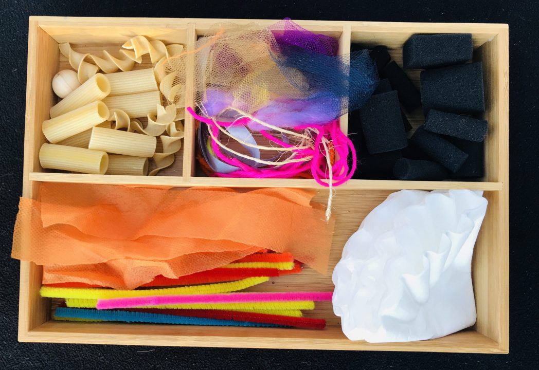 A divided wood tray with four compartments holds dry pasta, string, black foam pieces, pipe cleaners and coffee filters.