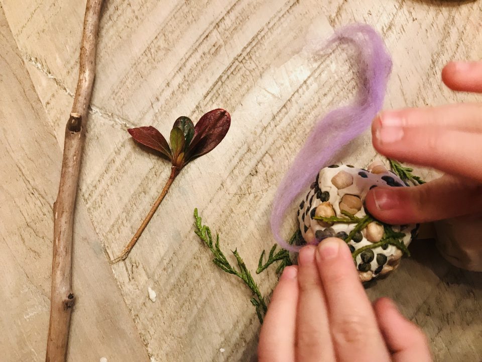 A child’s hands press dry beans and green lentils into a ball of clay. A stick, a red leaf, and purple wool are arranged nearby on the wood table.