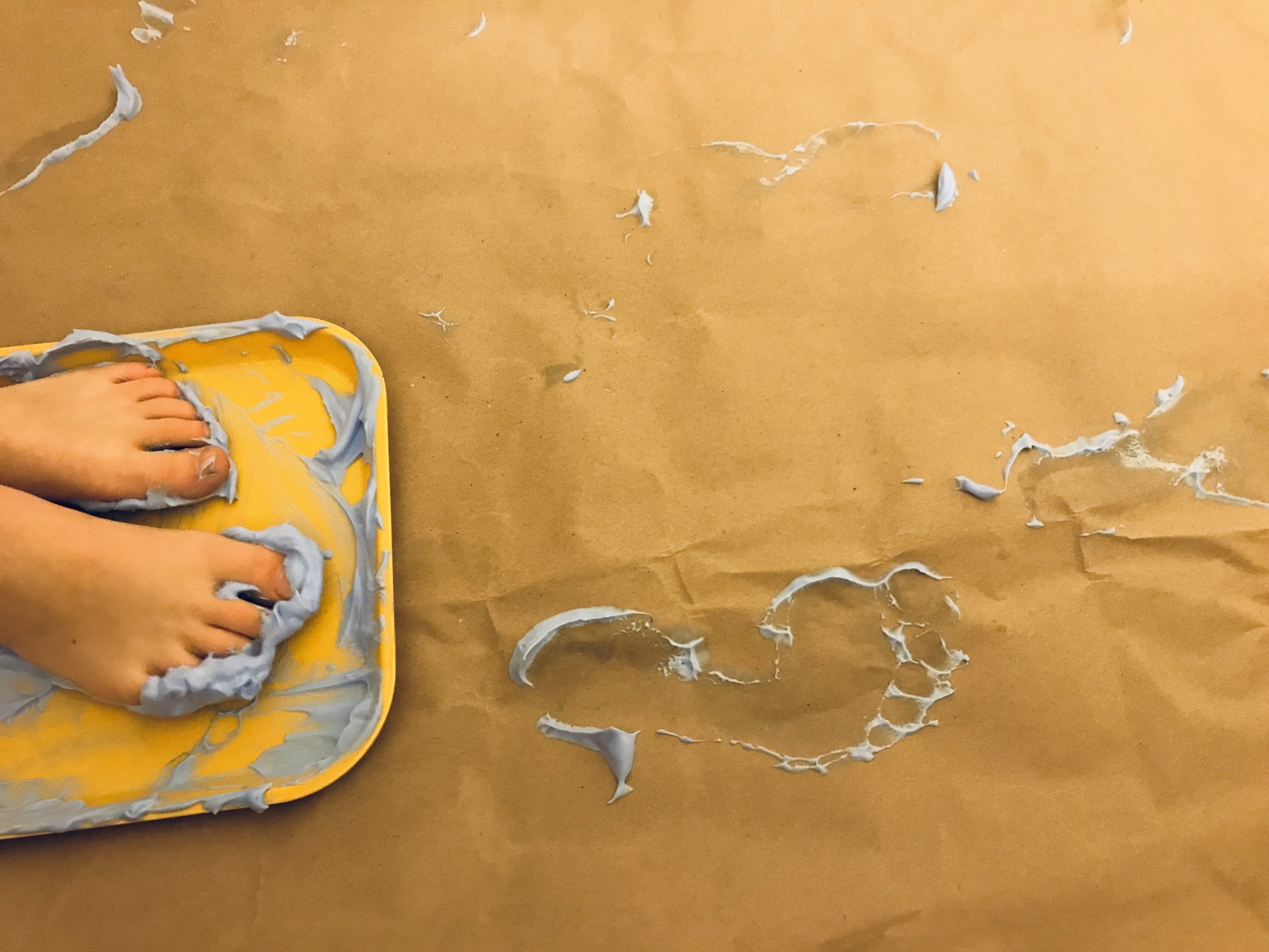 Two feet stand on a yellow tray of purple paint. Beneath the tray, three purple footprints make a circular pattern on the surface of brown paper.