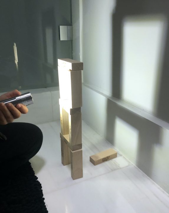 A tower of blocks casts a shadow on a wall. To the left, a hand points a flashlight at the tower. The shadow cast on the wall is much larger than the actual tower.