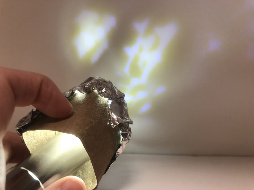 A hand holds a cardboard tube with a flashlight inside. The far end of the tube is covered in aluminum foil. On the wall behind the tube, a speckled pattern of light and shadow is cast.