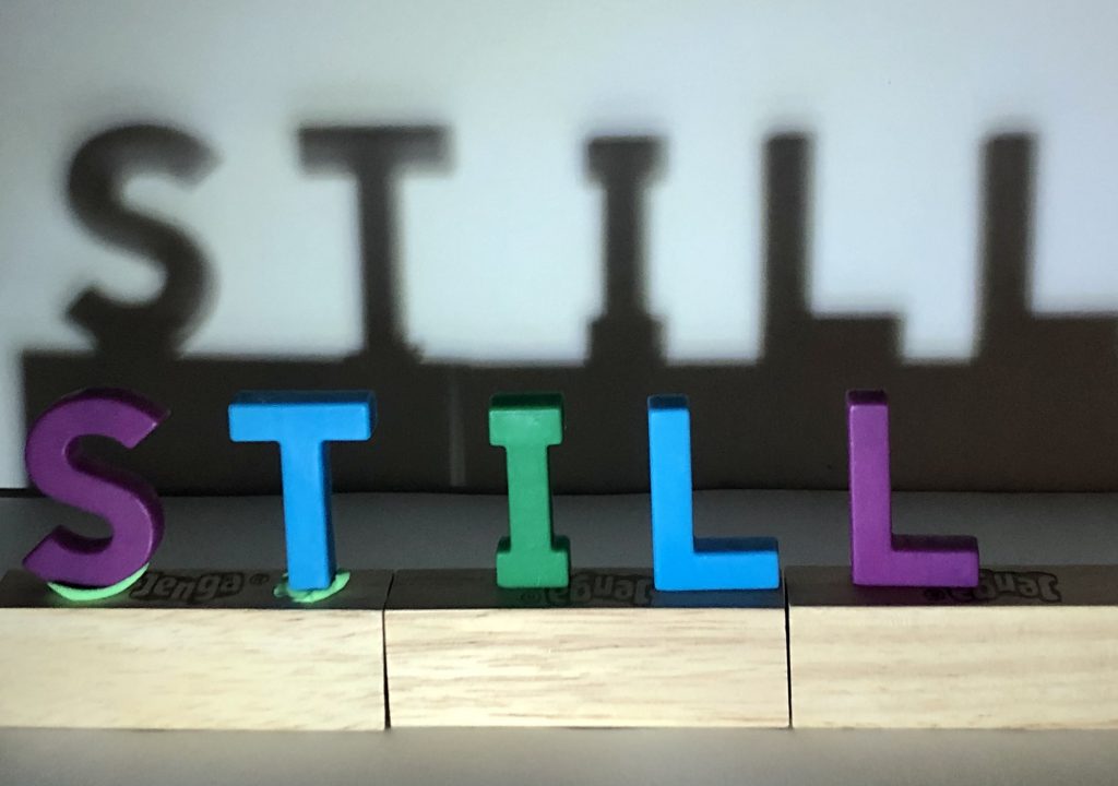 An arrangement of letter tiles spells the word “STILL.” On a wall behind it, the words are enlarged in a shadow.