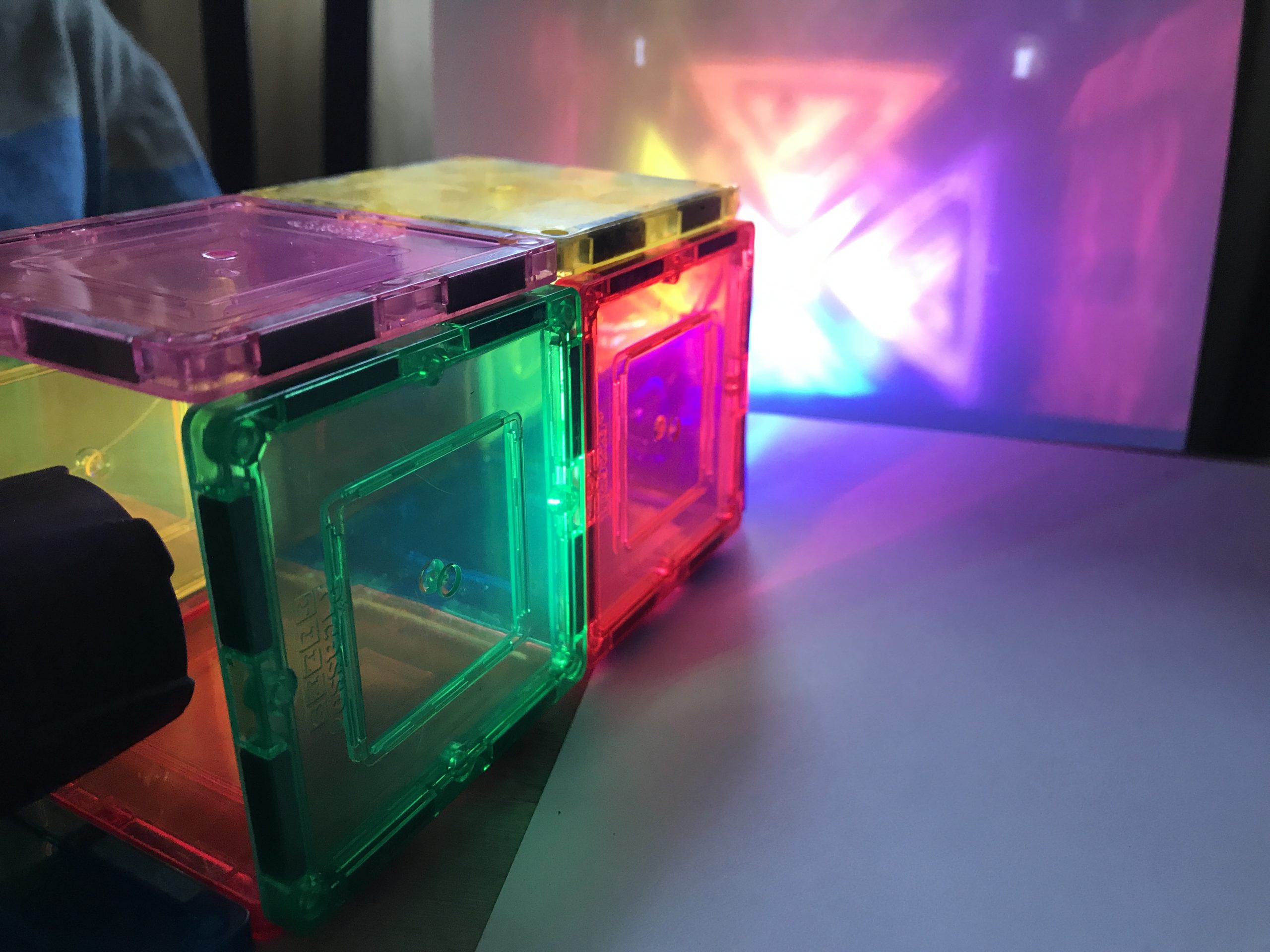 A flashlight shines through the end of a rectangular construction of colorful, translucent blocks, casting a projections of bright triangular shapes against a blank piece of paper.