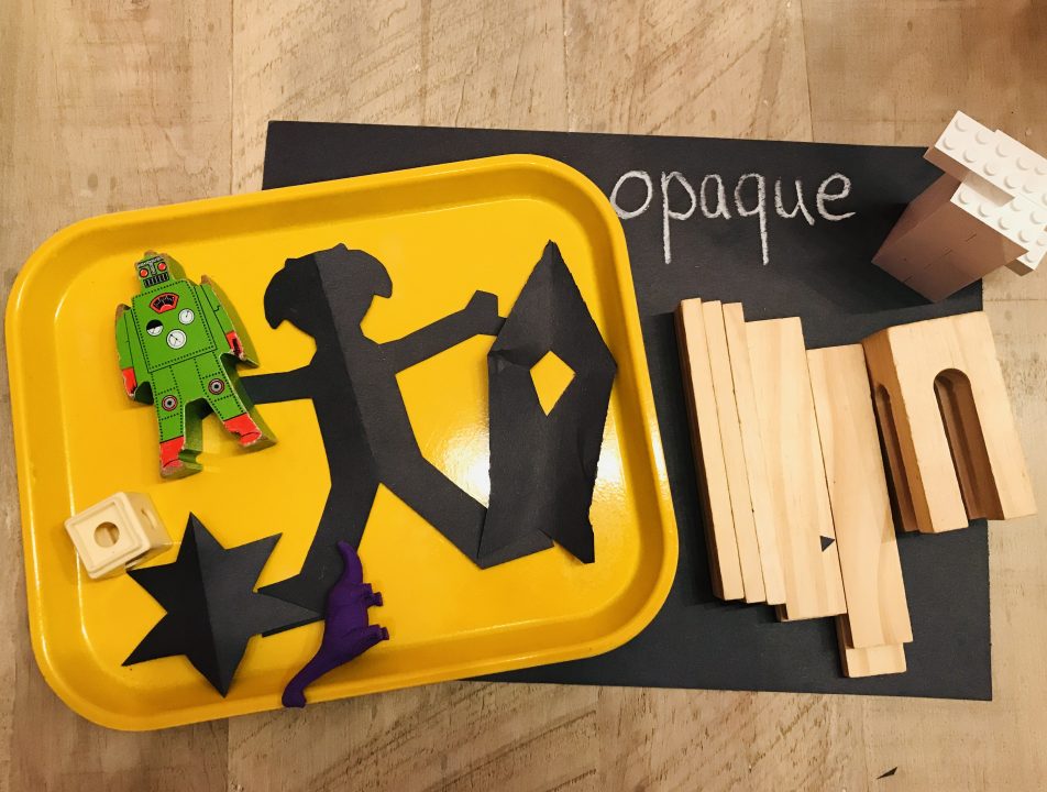 An arrangement of materials on a piece of black paper and a tray. The materials include paper cutouts, wood blocks, LEGO bricks, and a toy dinosaur. The paper reads “opaque.”