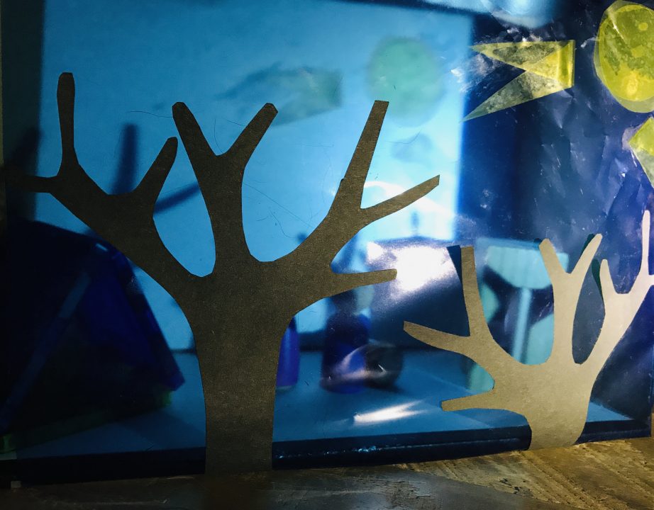 A shoebox shadowbox features a screen of translucent blue with black tree silhouette cutouts. Inside the box are small figures and a triangular structure. Shadows are cast on the back of the shoebox.