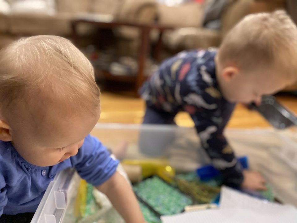 Two toddlers lean over a large bin filled with loose green, yellow, and blue materials. Each child has one hand inside the bin, and their faces look in toward the materials.