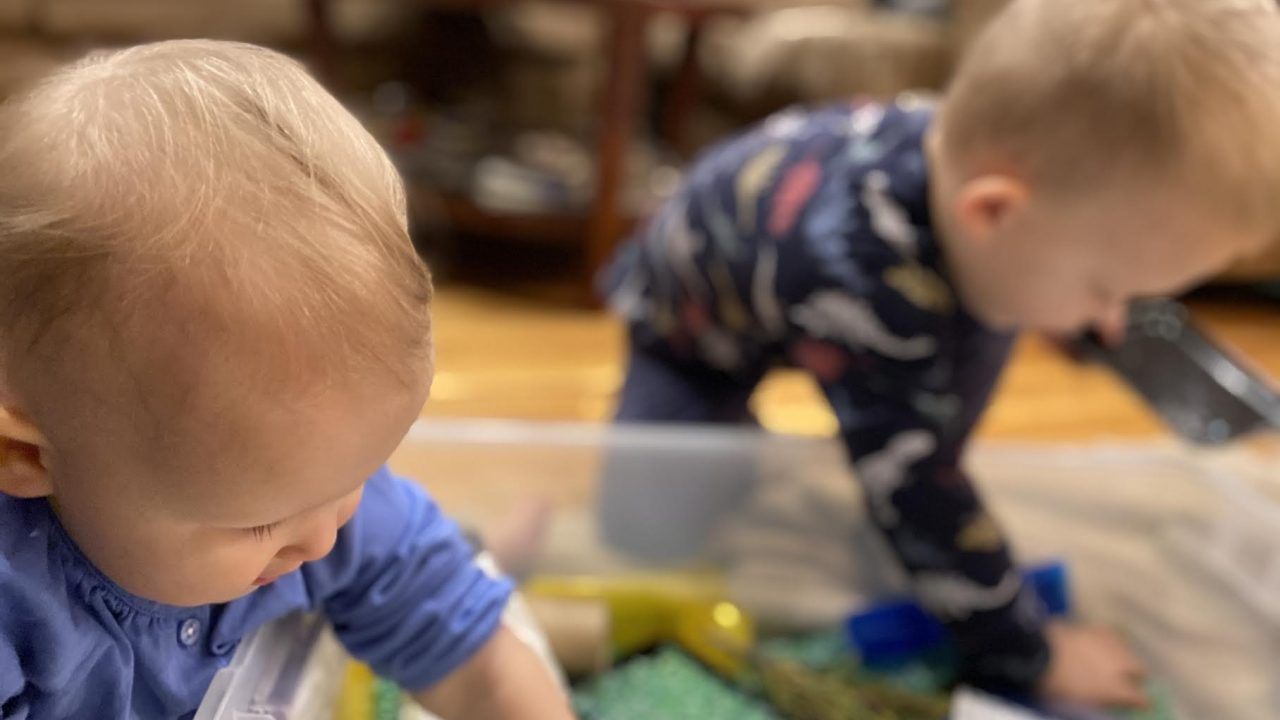 Two toddlers lean over a large bin filled with loose green, yellow, and blue materials. Each child has one hand inside the bin, and their faces look in toward the materials.