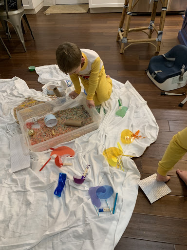 A boy looks inside a bin filled with multicolored materials. On a large white sheet surrounding him, materials are arranged in piles by rainbow color.