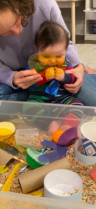 A baby sits on a woman’s lap in front of a sensory bin. The woman holds a yellow transparent circle in front of his face; the baby grasps a blue crinkly material.