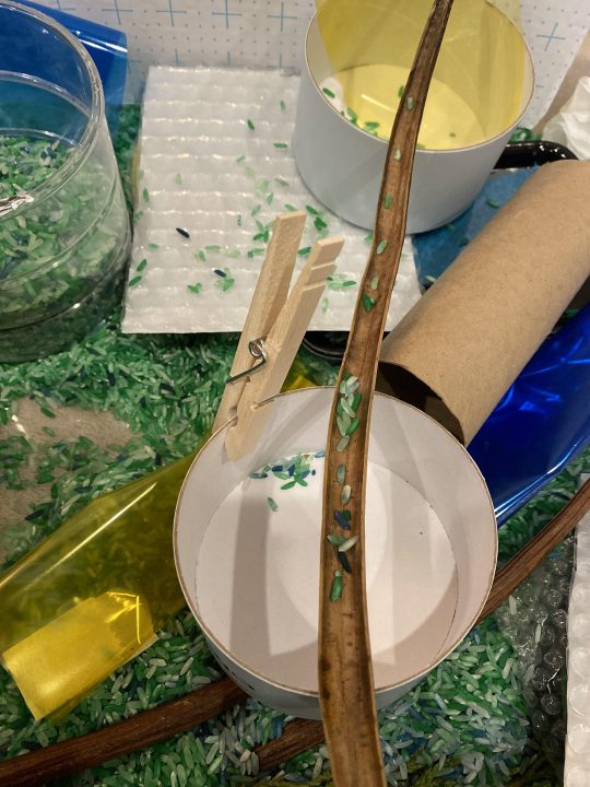 A long seed pod with a few grains of green rice is set atop two cup-shaped containers. Surrounding this structure are green rice and assorted loose materials.