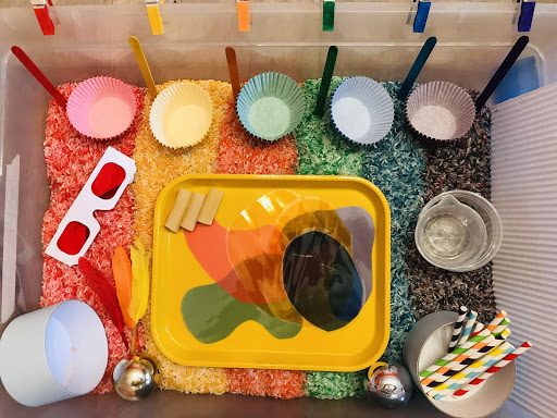 Bird’s-eye view of a sensory bin. Inside, rice and other materials are arranged in rainbow order. Materials include popsicle sticks, baking papers, feathers, and straws—all in multiple colors—as well as clear cups, and a yellow tray holding colorful shape cutouts.