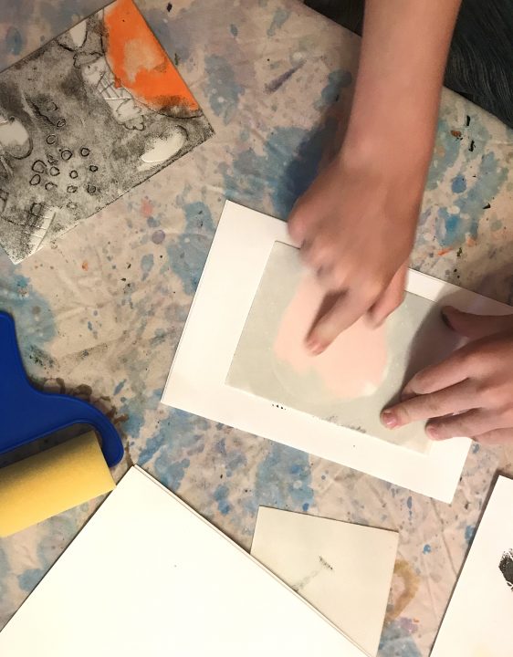 Two hands press styrofoam to paper; a black and orange design is slightly visible. A brayer and paper rest nearby.